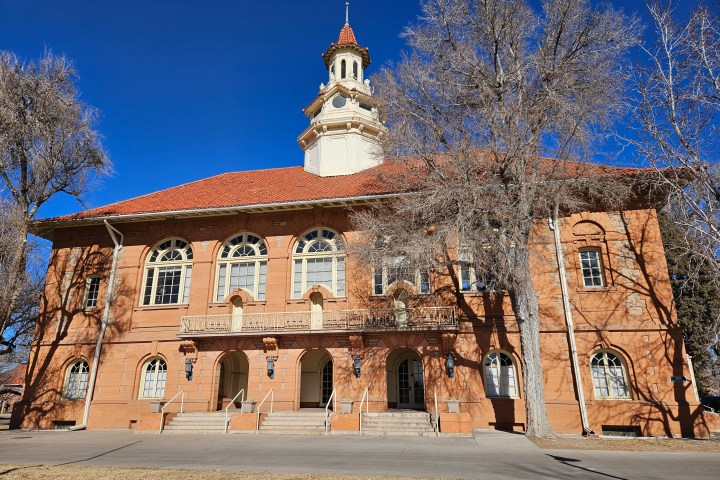 Historic brick building with a red roof and central tower in a sunny, tree-lined setting.
