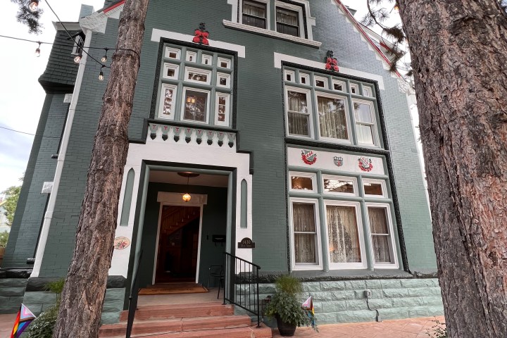 Historic green house with ornate trim and tall trees in front.