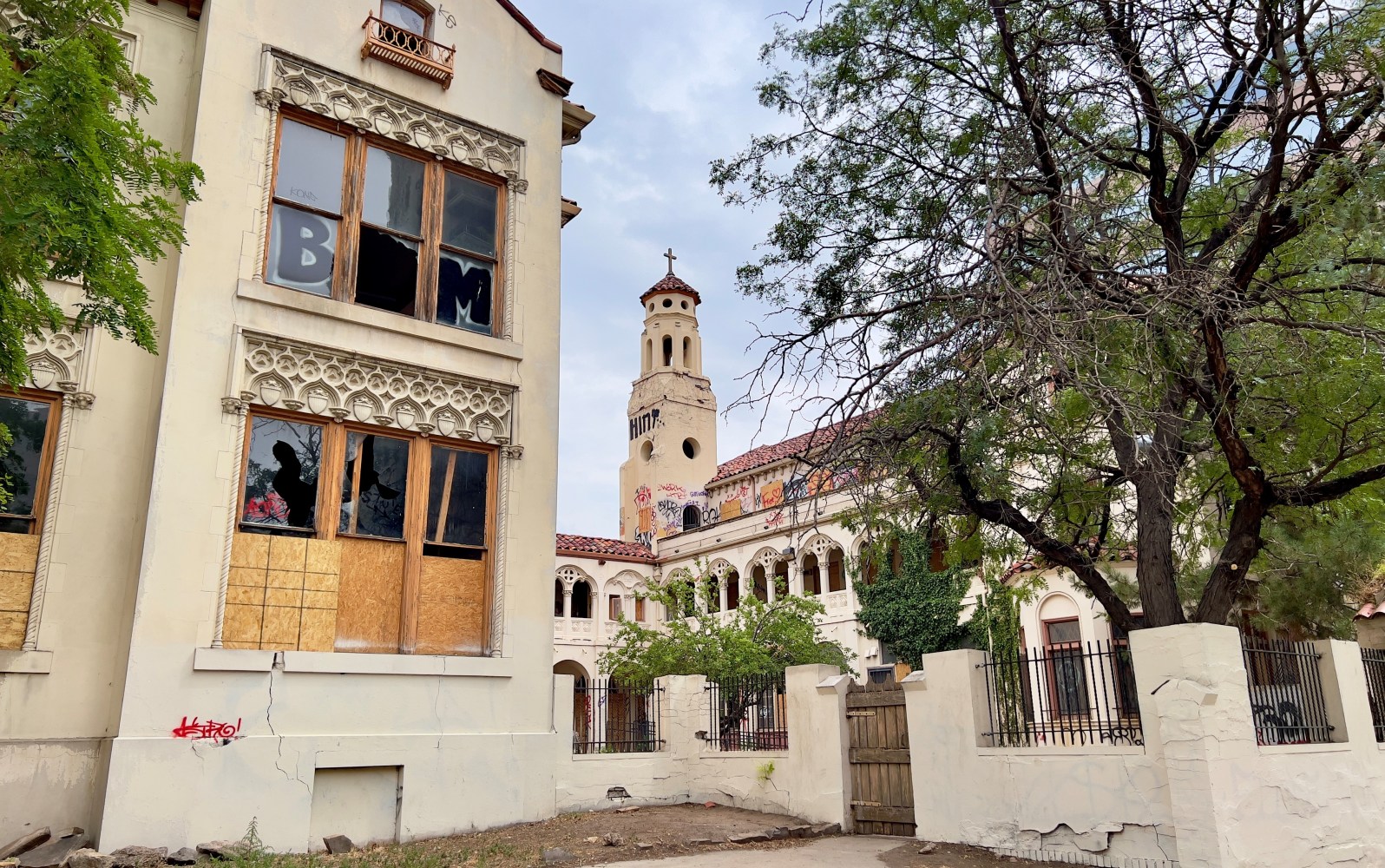 Historic building with boarded windows, graffiti, and a tower, surrounded by trees and a gated fence.
