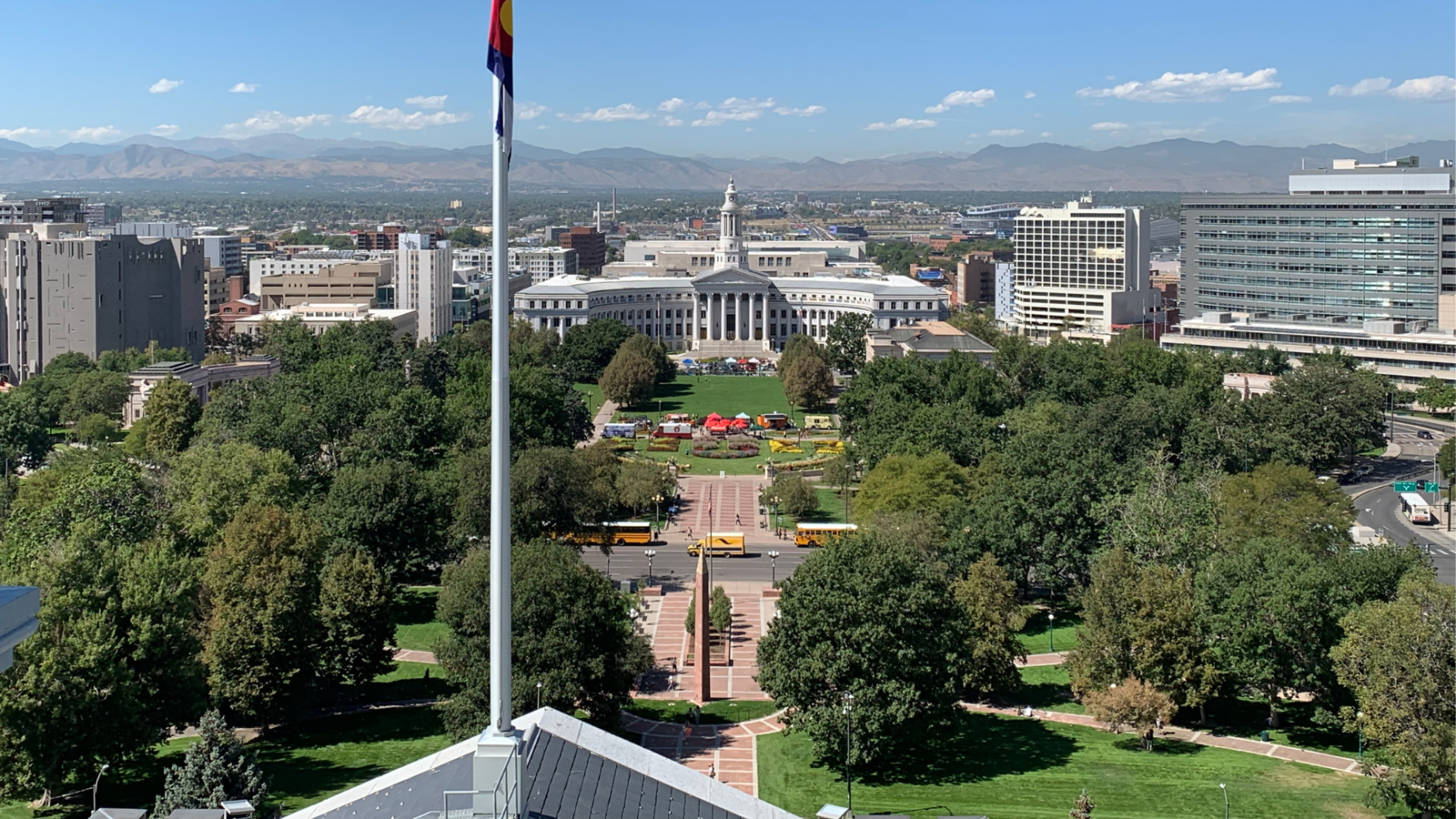 Aerial view of a city park with a flagpole, surrounded by trees, buildings, and mountains in the background.