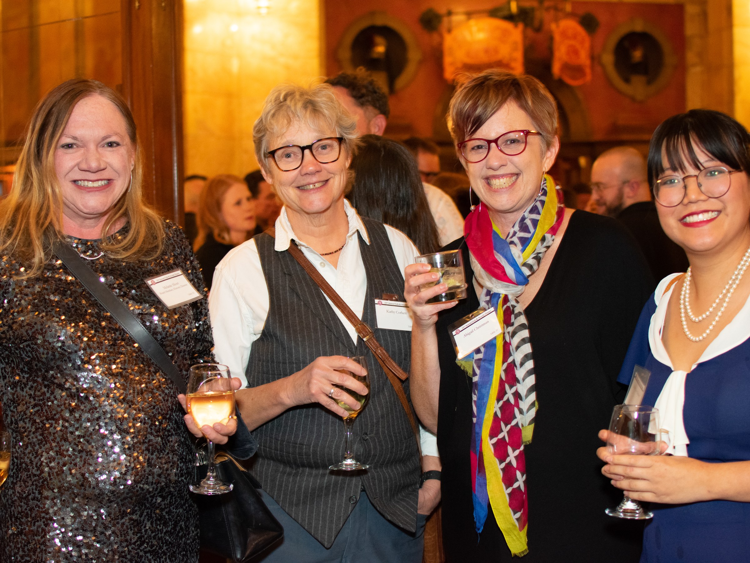 Four women smiling and holding drinks at an indoor event, wearing name tags.