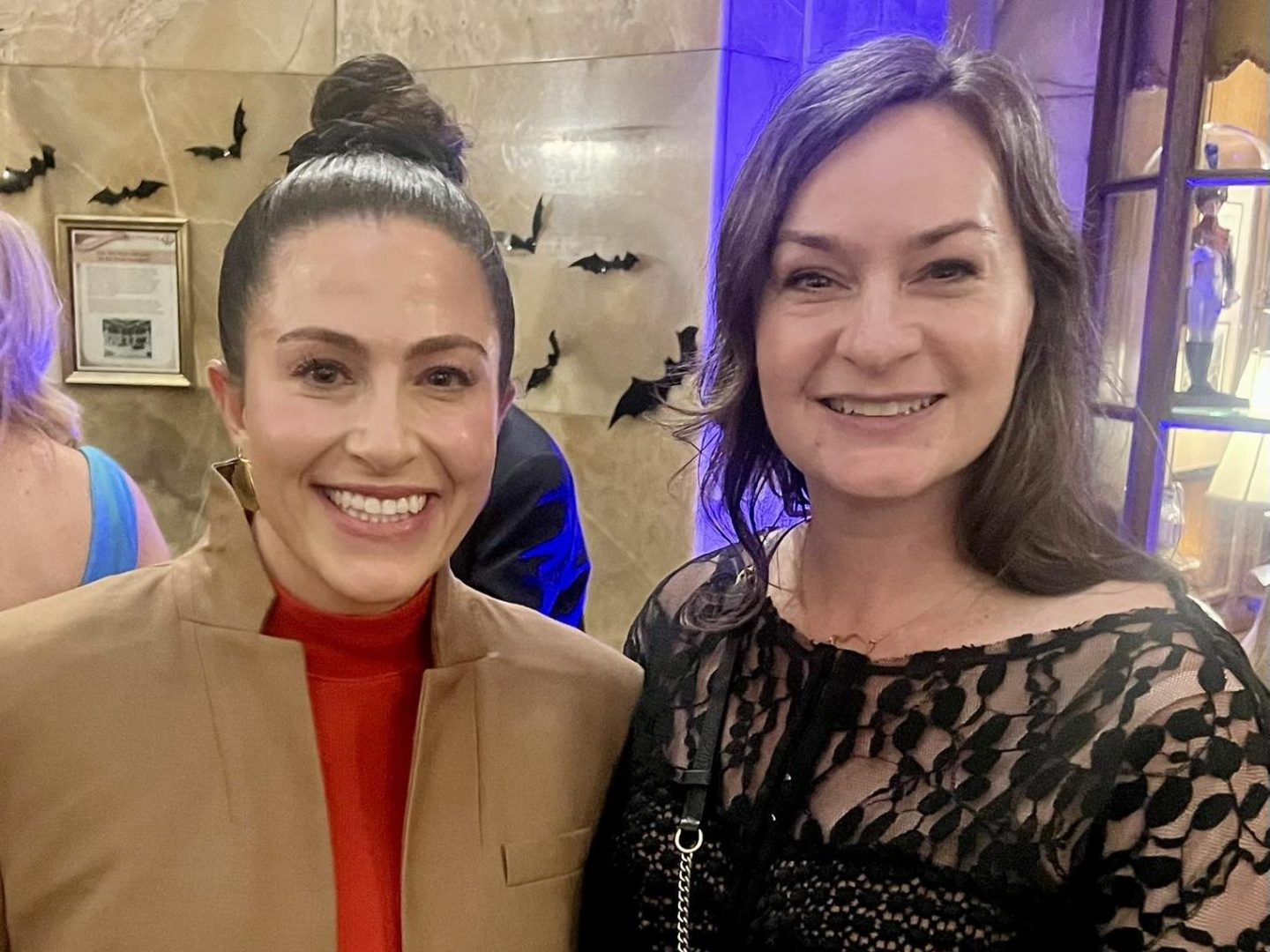 Two women smiling at event, one holding a wine glass, with bats decor on wall behind.