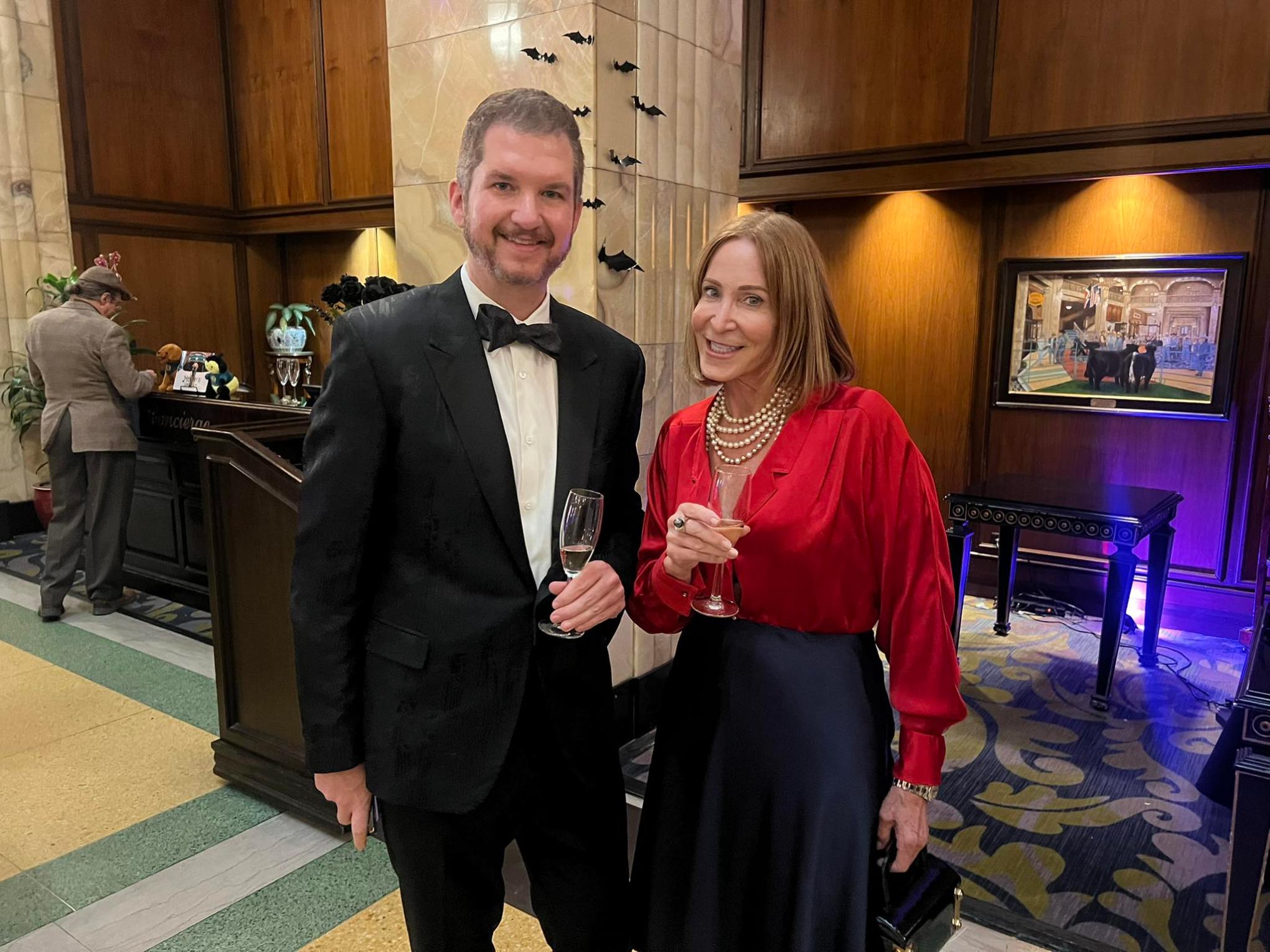 Man in tuxedo and woman in a red blouse holding drinks, standing in a stylish room.