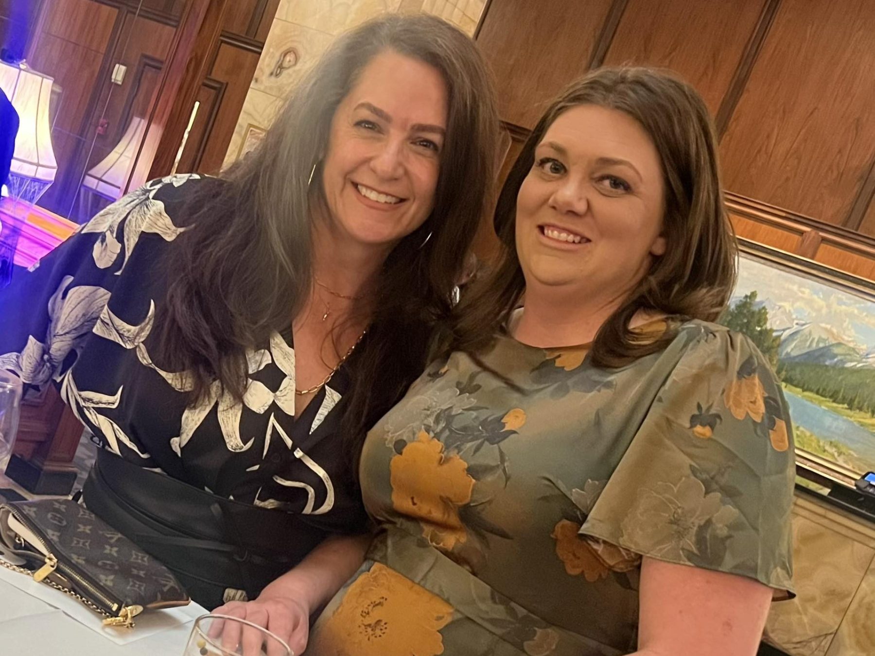 Two women smiling at an event, one holding a drink on a white table.
