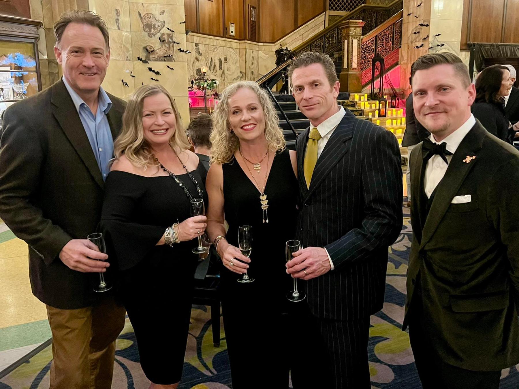 Five people dressed formally, smiling and holding champagne glasses at an indoor event.