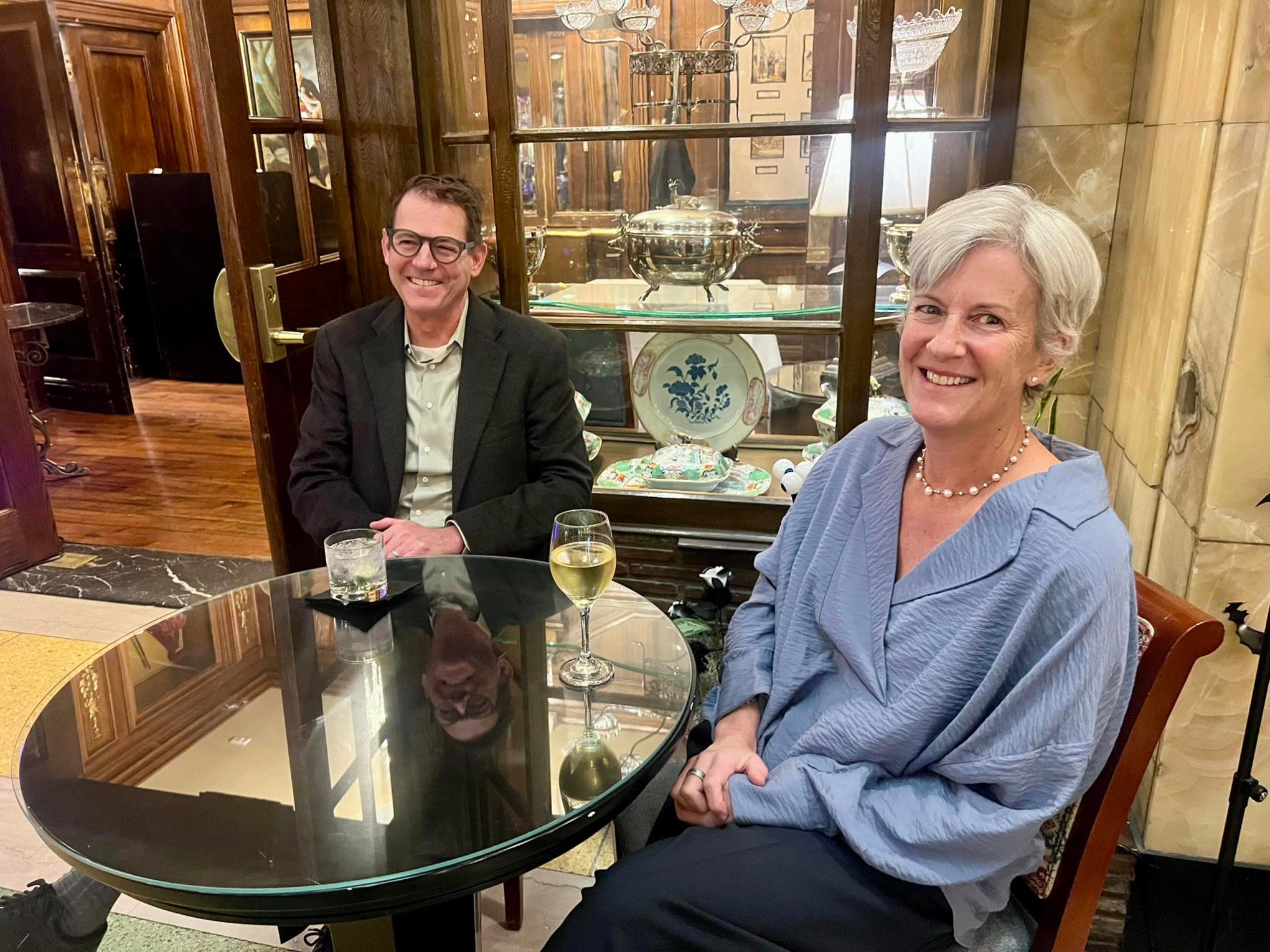 Two people seated at a round glass table with drinks in a cozy, elegant setting.