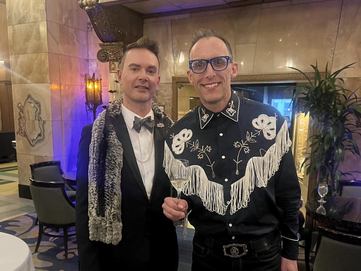 Two people in formal attire posing indoors, one in a suit with fur scarf, the other in a decorative shirt with glasses.