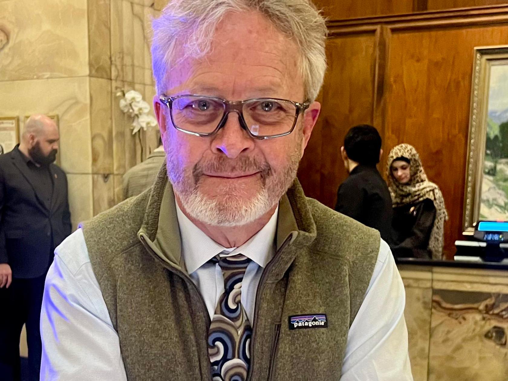 Smiling man with glasses, tie, and vest inside a wood-paneled room with people in the background.
