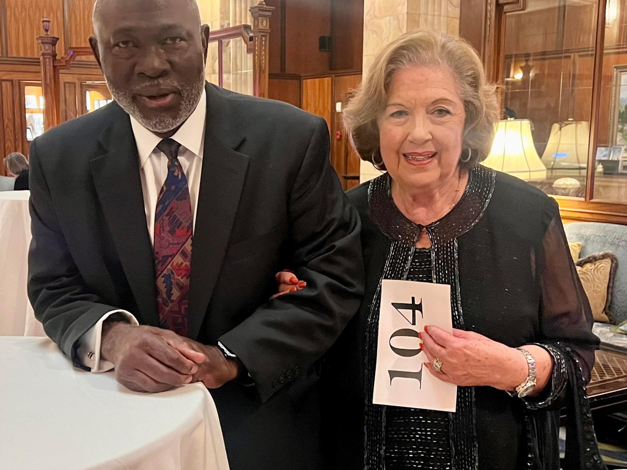 Two people in formal attire standing together indoors, one holding a sign with the number 104.