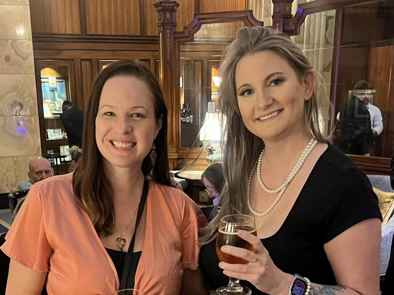 Two women smiling with drinks in a warmly lit room, wearing a peach dress and black dress with pearls.