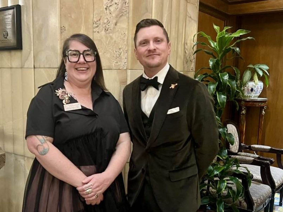 Two people dressed formally stand smiling in a decorated indoor setting with plants.