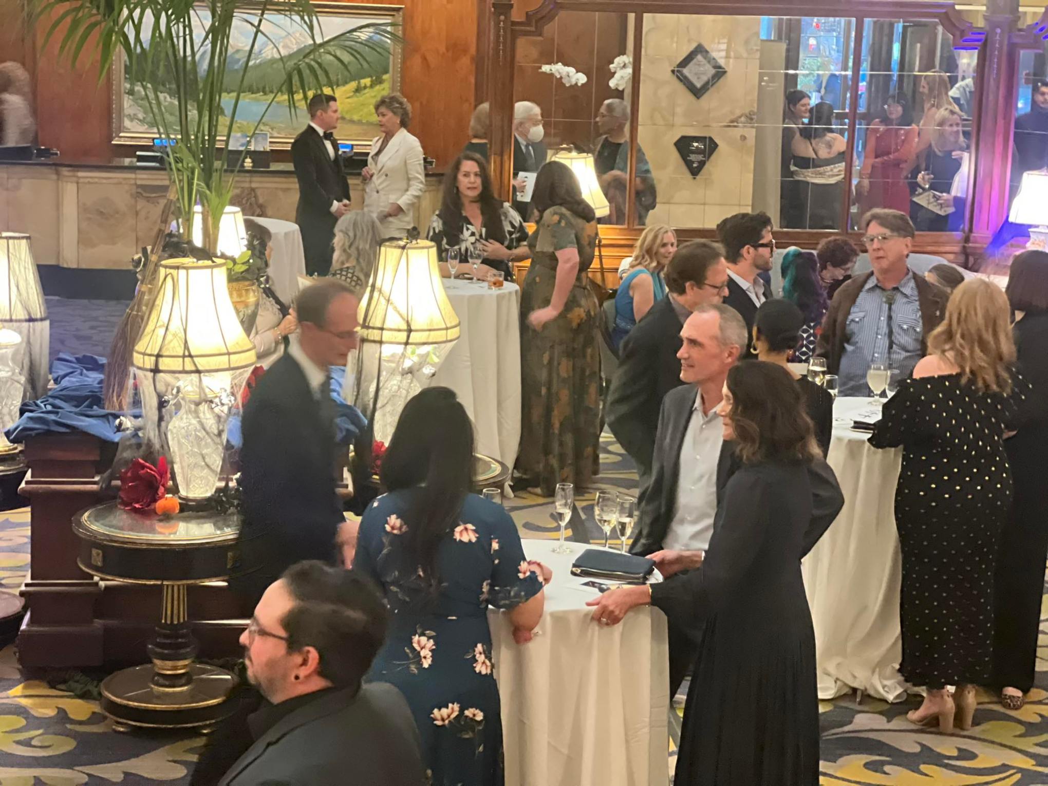 People in formal attire mingling at a reception with tables and lamps.