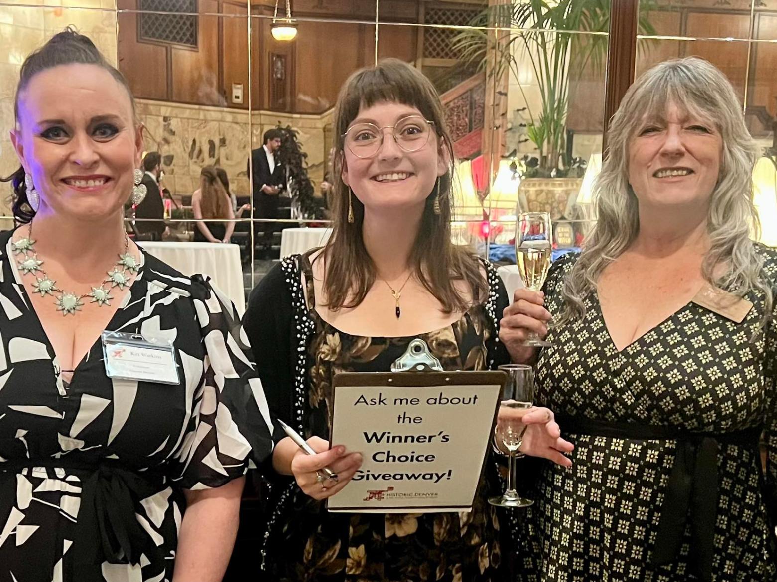 Three women smiling at an event, one holding a sign about a giveaway, with glasses of champagne.