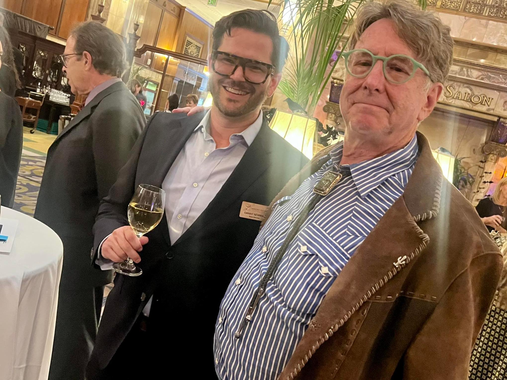 Two men in suits smiling at an indoor event, one holding a wine glass.