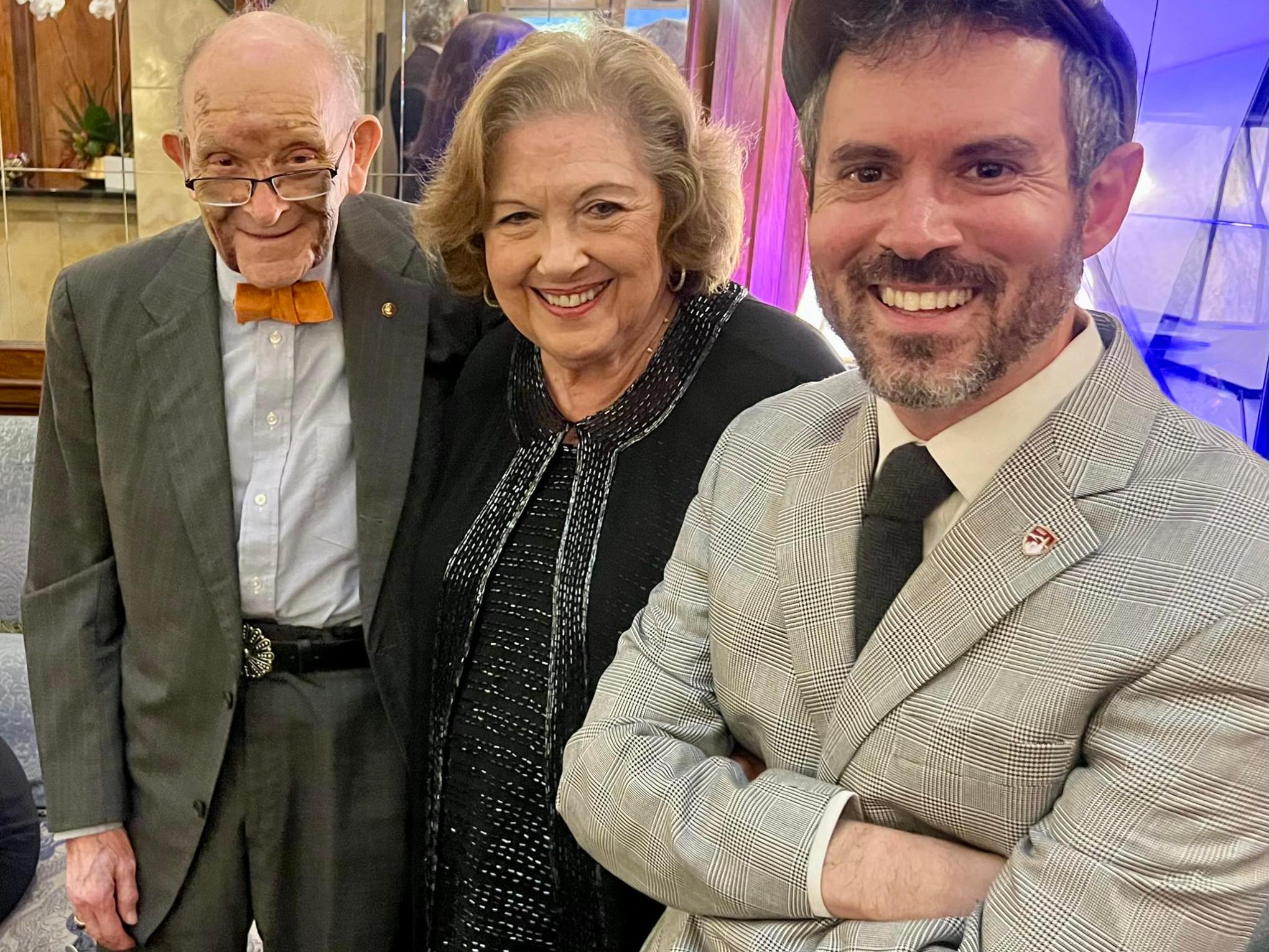 Three people smiling at an event; elderly man, middle-aged woman, and young man in a plaid suit and cap.