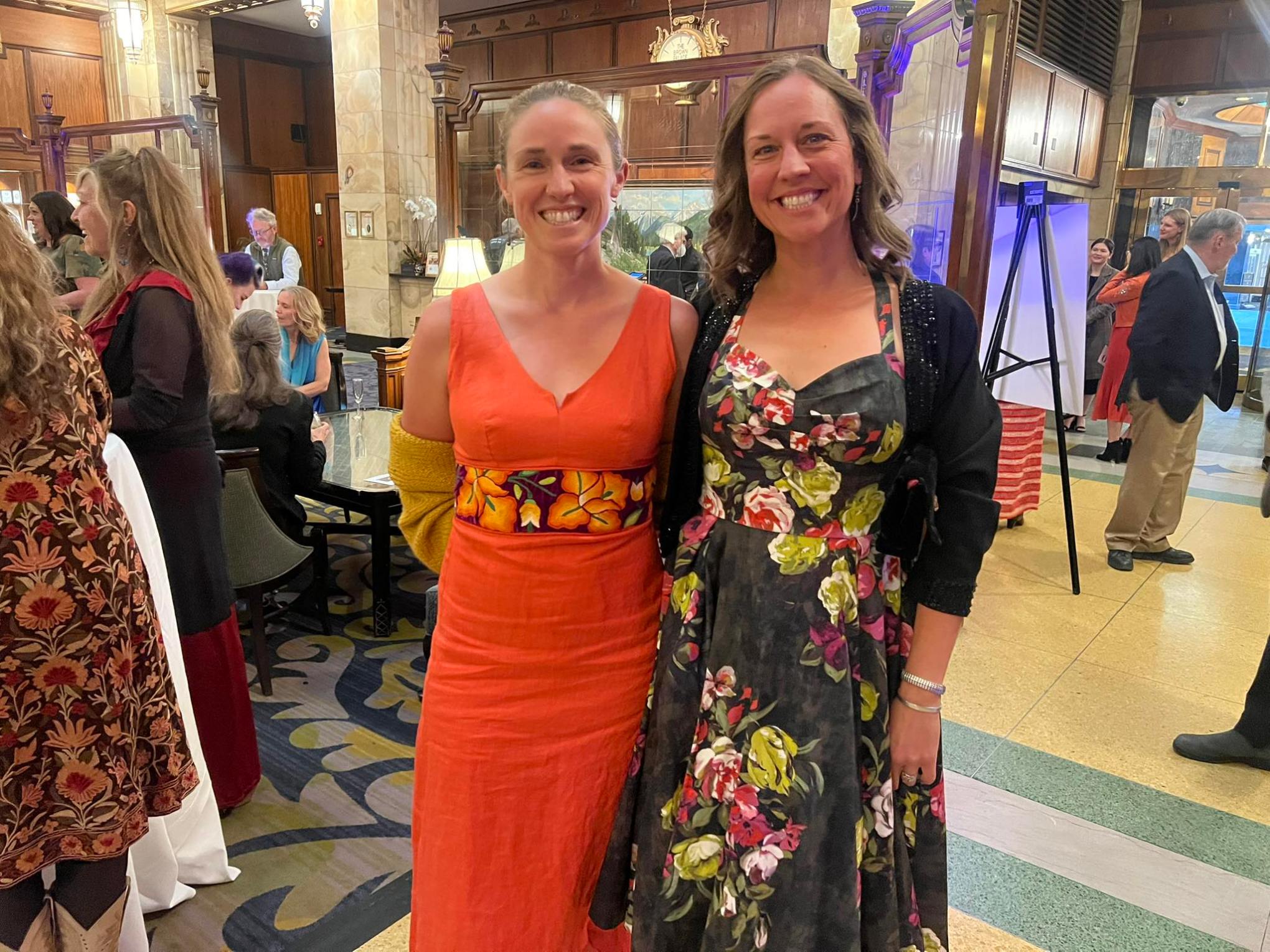 Two women in colorful dresses smiling at a formal indoor event with people in the background.