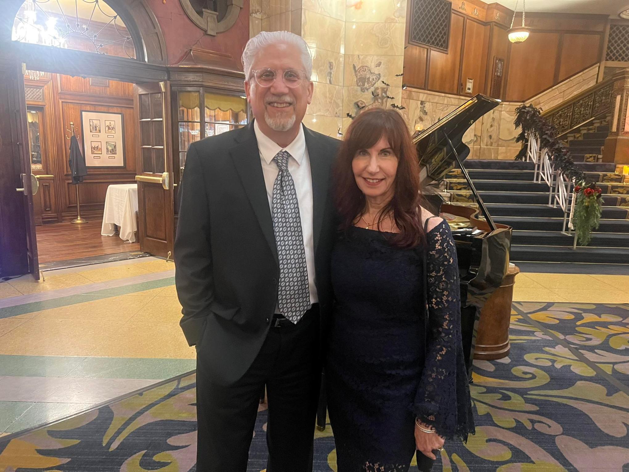 A man in a suit and a woman in a black lace dress standing in an elegant lobby with a grand piano in the background.