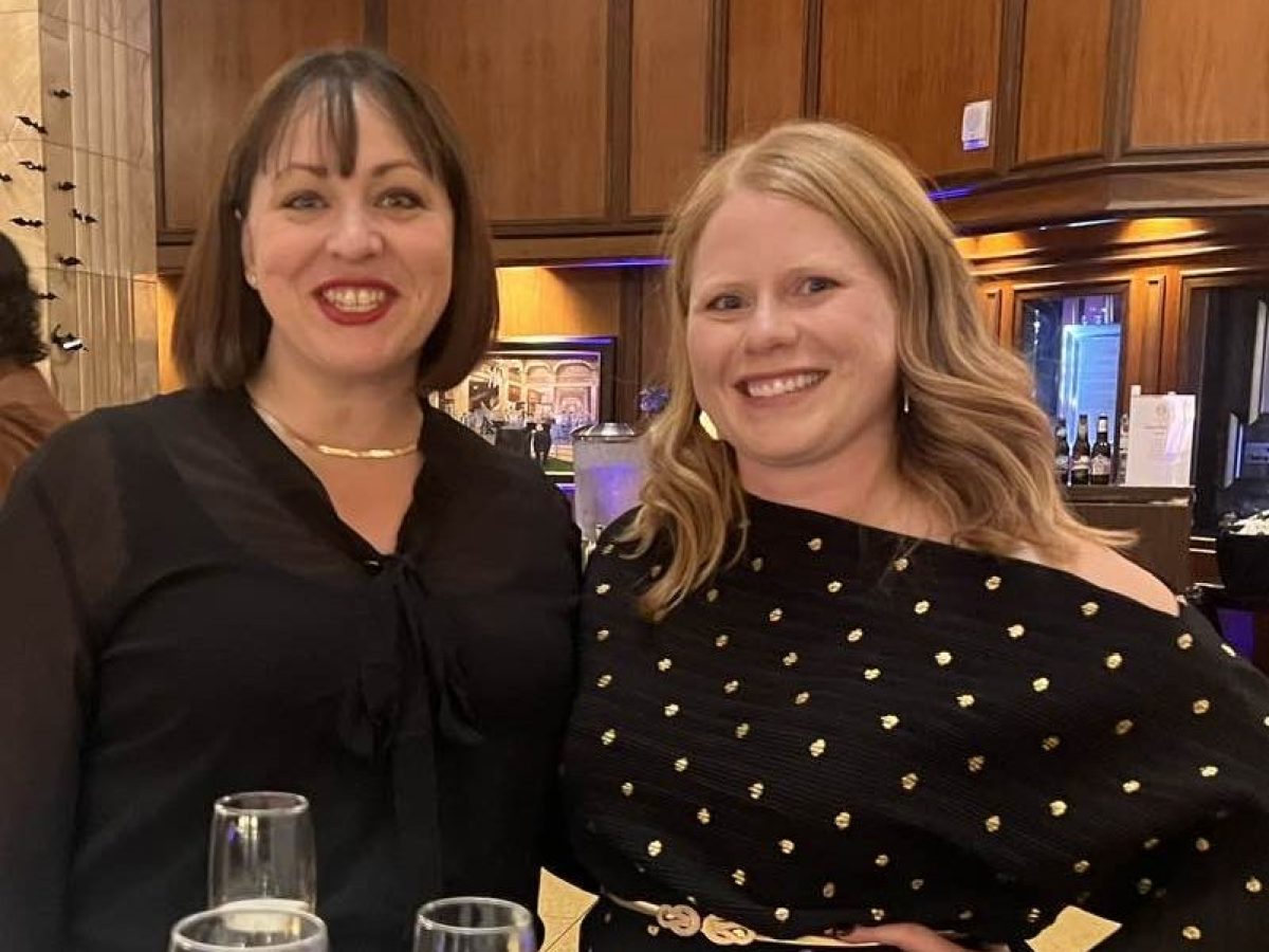 Two women smiling at a table with wine glasses in a wood-paneled room.
