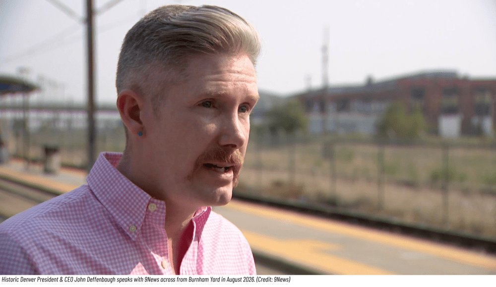 Person with mustache at train station, wearing a pink shirt.