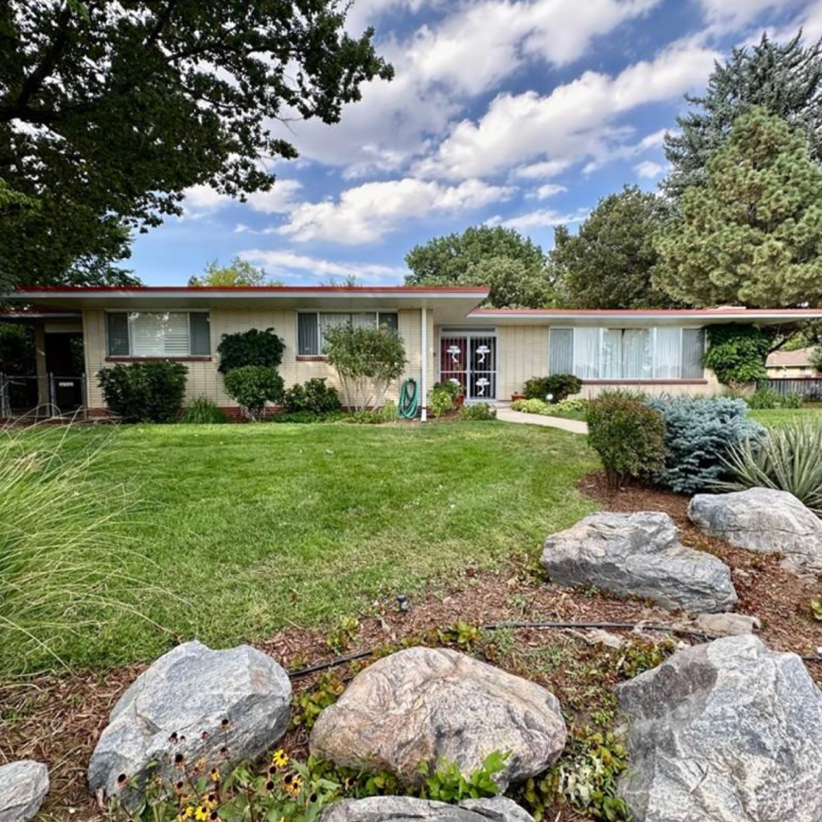 Single-story house with large front lawn, rocks, and trees.