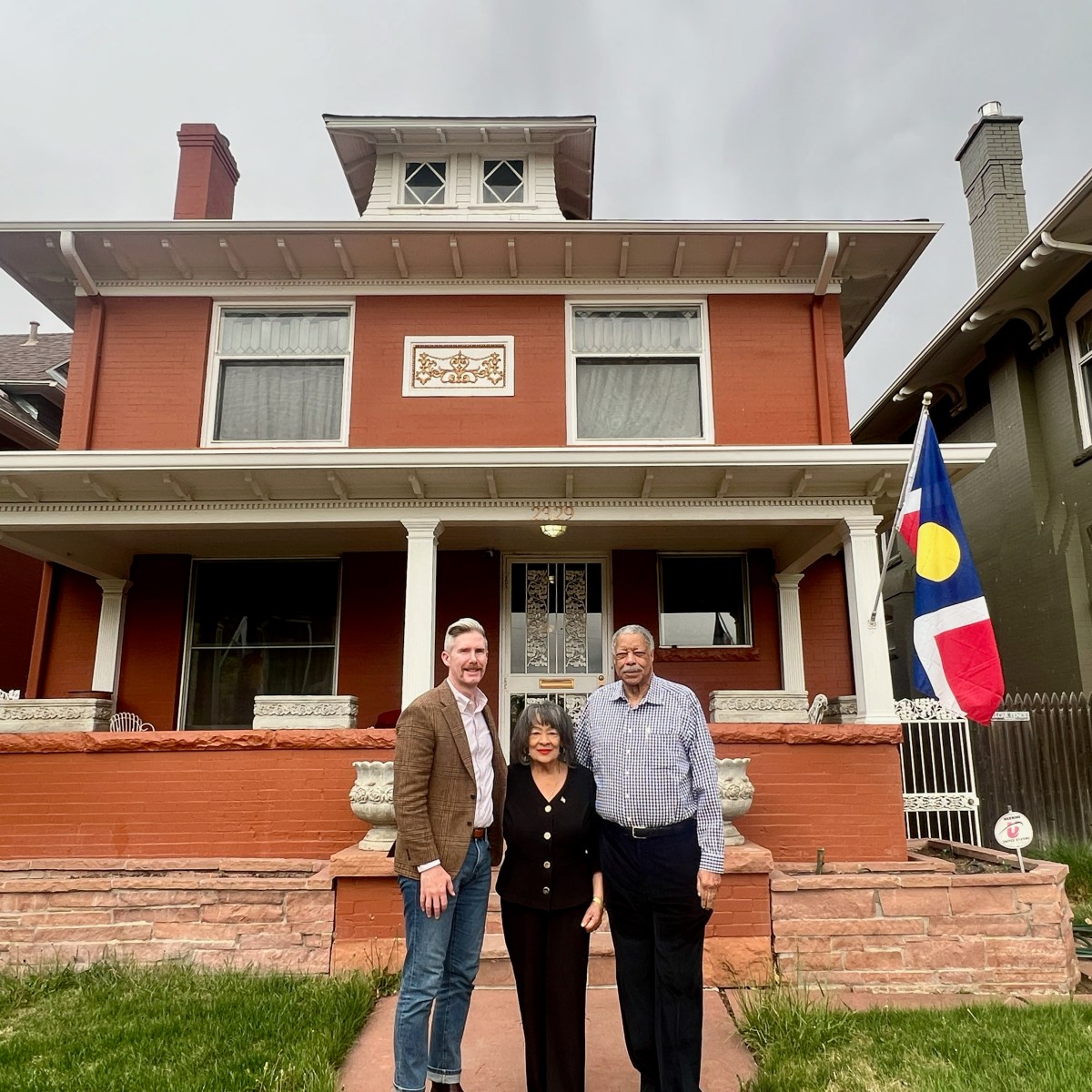 Three people stand in front of a red brick house with flags and a porch.