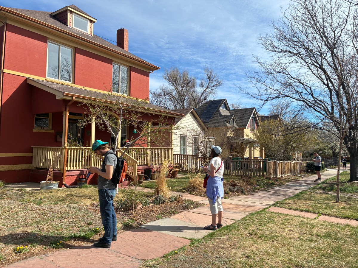 Two people with backpacks stand on a suburban sidewalk near colorful houses.